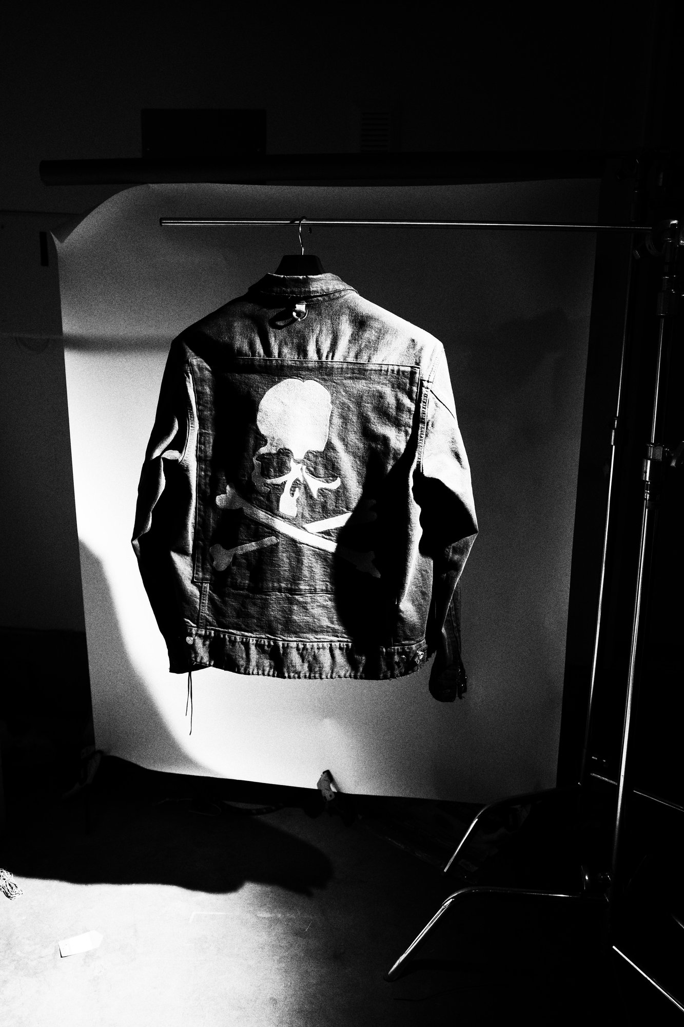 Black and white studio frame: the jacket on a hanger against a seamless backdrop under hard side light, stand and grip in shot.